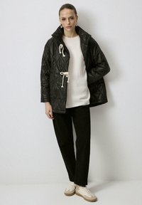 Chaqueta negra acolchada con un cuello alto, cerrada con cordón, llevada sobre un suéter crema. Combinada con pantalones negros y zapatillas beige.