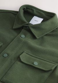 Veste vert foncé à col avec boutons-pression et poche à rabat, montrant un tissu texturé et une étiquette blanche avec le texte « NEXT » à l'intérieur du col.