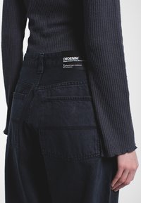 Mustat farkut korkealla vyötäröllä, joissa on näkyvä etiketti ja erottuva takataskun ompelu. Kangas on sileä.