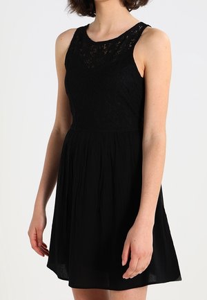 Hihaton musta mekko, jossa on pitsinen yläosa ja laskostettu hame, jota henkilö käyttää kädet rentoina sivuilla, yksiväristä taustaa vasten.