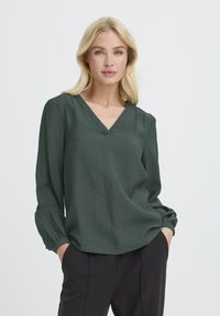 Femeie blondă cu machiaj ușor, purtând o bluză verde închis cu decolteu în V și mâneci lungi, împreună cu pantaloni negri, cu mâinile în buzunare, pe un fundal neutru.