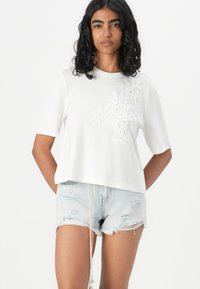T-shirt blanc à manches courtes avec broderie florale sur le côté gauche, associé à un short en jean déchiré bleu clair. Tissu doux, coupe décontractée.