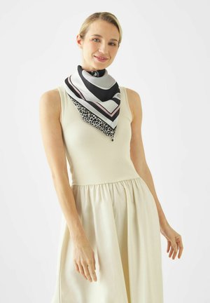 Vrouw draagt een mouwloze crèmekleurige jurk en een zwart-wit-roze sjaal met patroon, staand tegen een effen witte achtergrond.