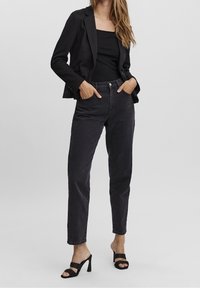 Černý blazer přes černý žebrovaný top, spárováno s černými džíny s vysokým pasem. Modelka má na sobě černé sandály na tenkém podpatku; neutrální pozadí.
