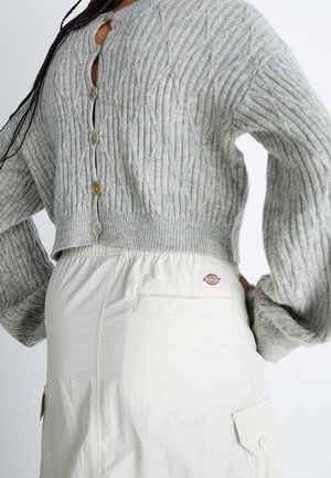 Personne portant un cardigan court en maille torsadée gris boutonné devant et une jupe crème taille haute avec poche arrière et écusson logo.