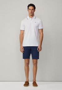 Polo bianca con rifiniture blu navy, abbinata a pantaloni corti blu navy. Il modello indossa scarpe scivolate marroni e si trova davanti a uno sfondo neutro.
