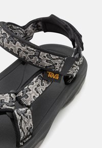 Sandály Teva s černou gumovou podrážkou, vzorovanými šedými a černými pásky a výraznou žlutou štítkem s logem.