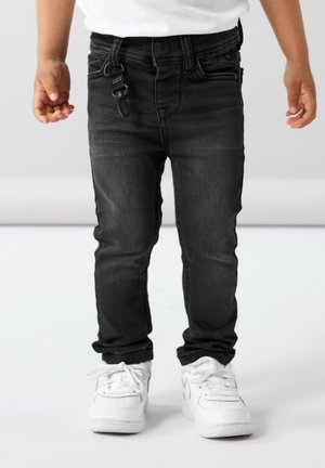Enfant debout portant un jean noir slim avec un porte-clés et des baskets blanches sur un fond clair uni.
