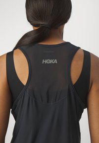 Čierny atletický tank top so priehľadnou zadnou panelou, širokými armovými otvormi a logom HOKA. Látka vyzerá ako ľahká a priliehavá, vhodná na šport.