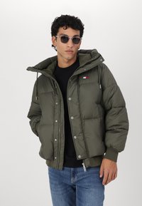 Chaqueta acolchada de color verde olivo con capucha desmontable, botones a presión y bolsillos laterales. Presenta un pequeño parche con logo en la parte superior izquierda del pecho.