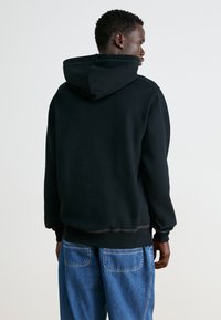 Felpa nera con cappuccio dal taglio comodo, con polsini e orlo a costine, e cuciture a contrasto. Indossata con pantaloni in denim blu chiaro.