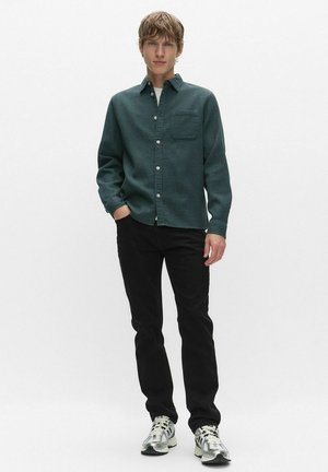 Ung man står med ena handen i fickan, iklädd mörkgrön skjorta med knappar, svarta byxor och vita sneakers mot en enfärgad vit bakgrund.