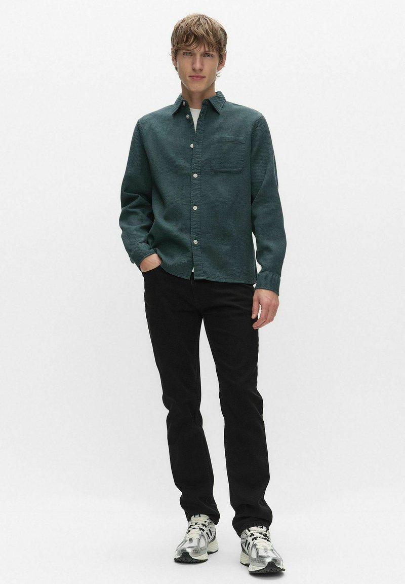 Jeune homme debout, une main dans la poche, portant une chemise boutonnée vert foncé, un pantalon noir et des baskets blanches, sur un fond blanc uni.