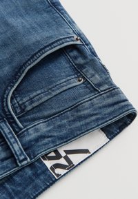 Jean en denim bleu plié avec poche avant visible, rivets et une étiquette de taille blanche marquée "27" sur fond blanc.