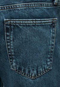 Κοντινό πλάνο πίσω τσέπης μπλε τζιν με ορατή πορτοκαλί ραφή σε υφασμένο ύφασμα.