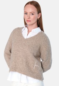 Beige v-hals genser laget av mykt stoff, med en avslappet passform, ribbede mansjetter, og en liten brodert "H" detalj på fronten.