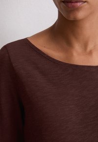 Brązowa, fakturowana bluzka z szerokim dekoltem w łódkę, z prążkowanym wykończeniem i minimalnymi detalami przeszycia. Odpowiednia do noszenia na co dzień.