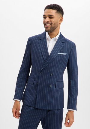 Mann trägt einen marineblauen Nadelstreifen-Zweireiher-Anzug mit weißem Hemd, lächelt und schaut seitlich vor einem einfarbigen Hintergrund.