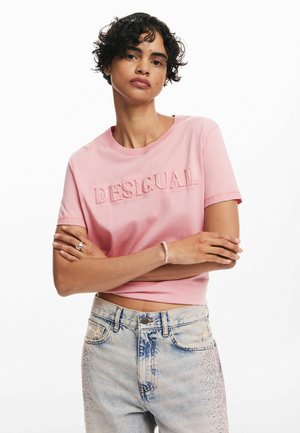 Light pink cotton t-shirt featuring embossed "DESIGUAL" text. Round neckline, short sleeves, paired with faded blue jeans with embellishments.