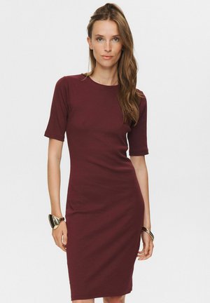 Woman with long brown hair wearing a fitted burgundy knee-length dress and metallic bracelets on both wrists, standing against a white background.