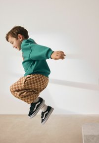 Băiat în pulover teal, pantaloni bej în carouri și adidași negri cu talpă albă, sărind în aer pe un fundal deschis.