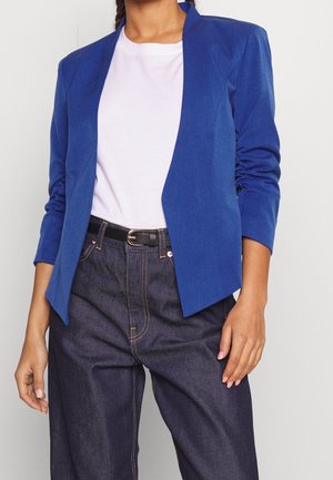 Femme portant un blazer bleu roi ouvert sur le devant, un t-shirt blanc, un jean en denim foncé, et une ceinture noire avec une boucle dorée.