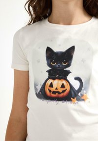 Valkoinen lyhythihainen t-paita, jossa on mustan kissan grafiikka viittassa istumassa kaiverretun kurpitsan päällä, jossa on hymyilevä naama ja tähdenkoristeluja.