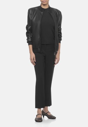 Donna che indossa un bomber in pelle nero, top nero, pantaloni neri corti e scarpe nere a tacco basso con motivo, in piedi su sfondo bianco.