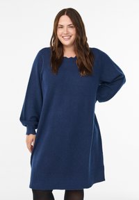 Femme souriante avec de longs cheveux bruns, portant une robe pull bleu marine ample à manches ballon et des collants noirs sur un fond blanc.