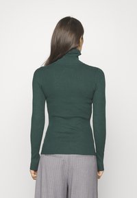 Pull à col roulé côtelé vert foncé avec des manches longues, au design ajusté et à la texture subtile. Porté avec un pantalon gris à rayures.