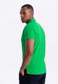 Zelené polo tričko vyrobené z bavlny, má klasický límec, krátké rukávy a boční rozparky. Nosí ho mužský model, střih je slim fit.