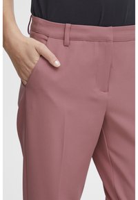 Rose-colored tailored pants with a smooth texture, featuring a side pocket and a mid-rise waistband. Close-up view highlights fit and fabric.