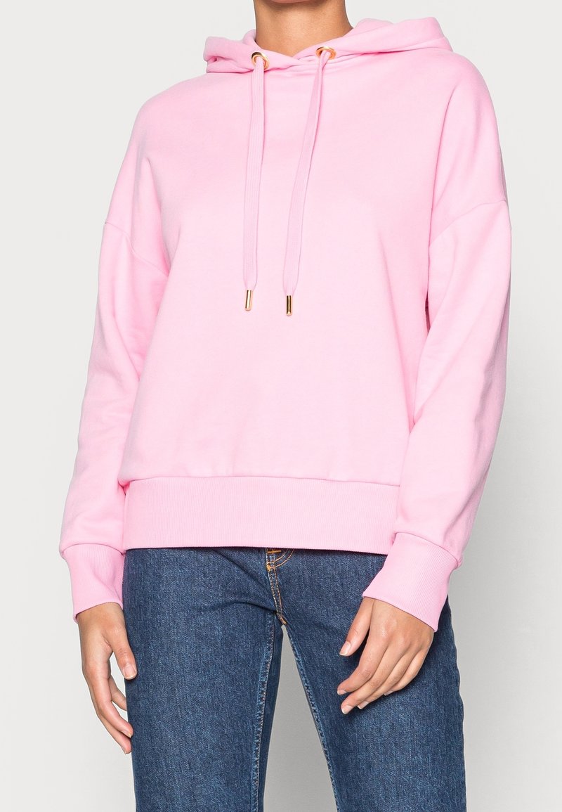 Sudadera con capucha de color rosa claro con cordones y detalles dorados, presenta un ajuste holgado y puños acanalados, combinada con vaqueros oscuros.
