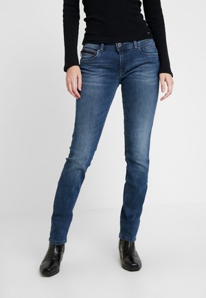 Jeans ajustados de tiro bajo, azul oscuro, con un lavado desgastado, cinco bolsillos y cierre de cremallera. El modelo lo combina con una blusa negra de manga larga y botas negras.