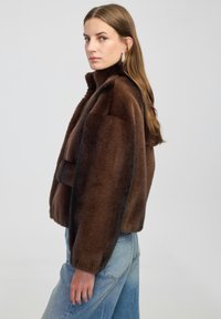 Braune Kunstpelzjacke mit hohem Kragen, kastigem Design und tief geschnittenen Schultern. Getragen über hellblauen Denim-Jeans.