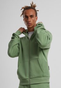Haut en sweat à capuche zippé vert en tissu doux, avec deux poches avant, des poignets et un ourlet côtelés, et un branding subtil sur la poitrine.