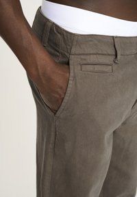 Grå bukser med en glat tekstur, der har en flad front, sidelommer og et hvidt taljebånd, der delvist er synligt øverst.