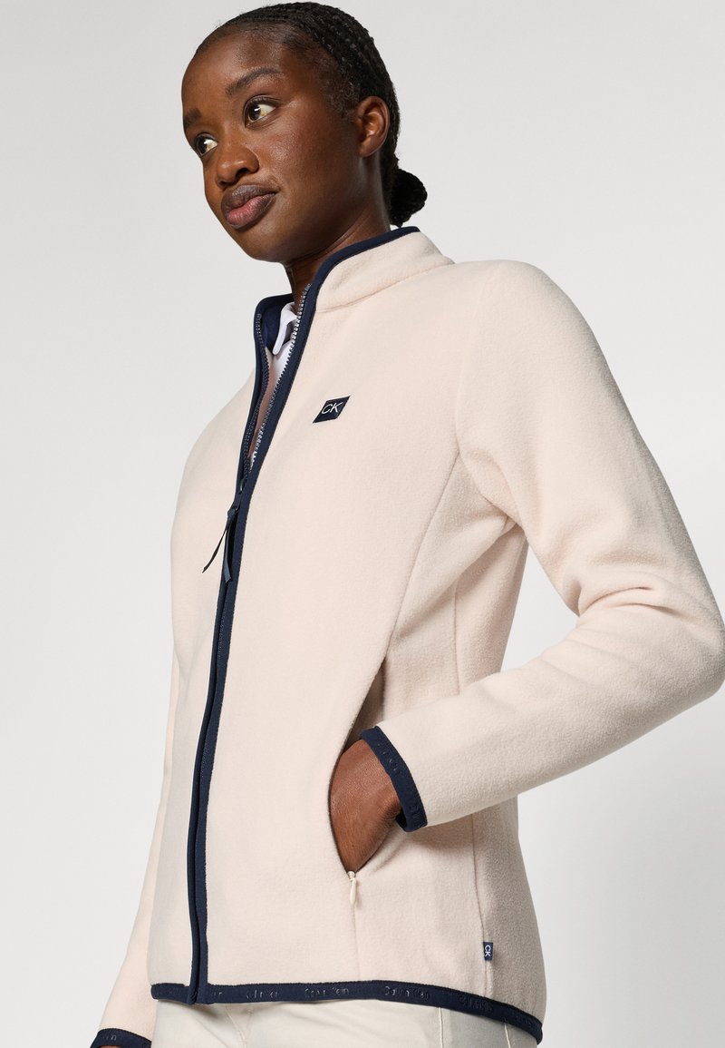 Helles rosa Fleece-Jacke mit einem marineblauen Reißverschluss und Bündchen, ausgestattet mit Seitentaschen und einem kleinen Logo auf der Brust. Weiche Textur.