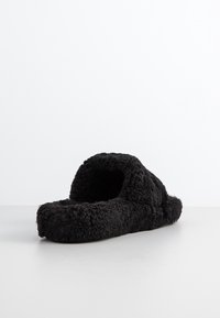 Chausson noir duveteux à ouverture arrière avec semelle épaisse, conçu pour le confort et la chaleur à l'intérieur, présenté sur une surface blanche.
