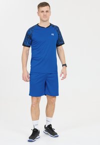 Blå idrottströja med svarta mönstrade ärmar, i kombination med blå shorts. Bärs med svarta sneakers och vita strumpor, med loggdetaljer.