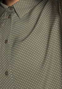 Green, short-sleeve shirt with a polka dot pattern; features a button-down collar and brown buttons; smooth fabric texture.