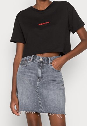 Czarny krótki T-shirt z czerwonym napisem „DOLCE VITA” w połączeniu z jasnoniebieską, wysokim stanem spódnicą jeansową z surowym wykończeniem i ręką w kieszeni.