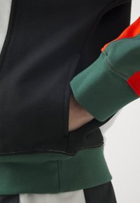 Main dans la poche d'un sweat-shirt noir avec manches à blocs de couleurs vertes et rouges et ourlet côtelé vert.