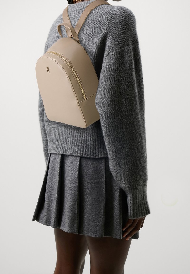Personne portant un sac à dos beige sur un pull en maille gris et une jupe plissée grise, vue de dos sur un fond uni.