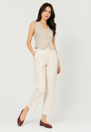 Woman with long dark hair wearing beige sleeveless buttoned vest, high-waisted cream pants, and brown flat shoes, standing with hands in pockets.