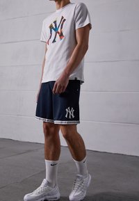 T-shirt blanc avec un logo "NY" multicolore, associé à un short marine avec un emblème "NY" blanc et des détails gris, complété par des baskets blanches.
