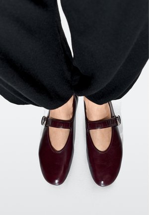 Bordeauxrode leren Mary Jane schoenen met een ronde neus, een uitsnijdingsdetail en een verstelbare gespsluiting. Draagt ze met zwarte broek.