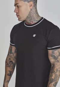 Juodas marškinėliai su balta apdaila, apvalia iškirpte, prigludusio dizaino ir mažas logotipas ant krūtinės; papuošti detalizuotais tatuiruotėmis ant rankos.