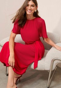 Femme souriante avec de longs cheveux bruns portant une robe rouge et des escarpins rouges, assise sur un fauteuil en tissu texturé avec des pieds en métal.