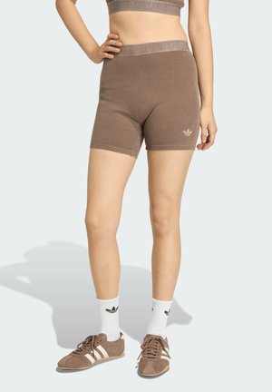 Persona che indossa pantaloncini marroni a coste e top corto abbinato, calzini bianchi Adidas e scarpe da ginnastica marroni Adidas, in piedi con la mano sul fianco.